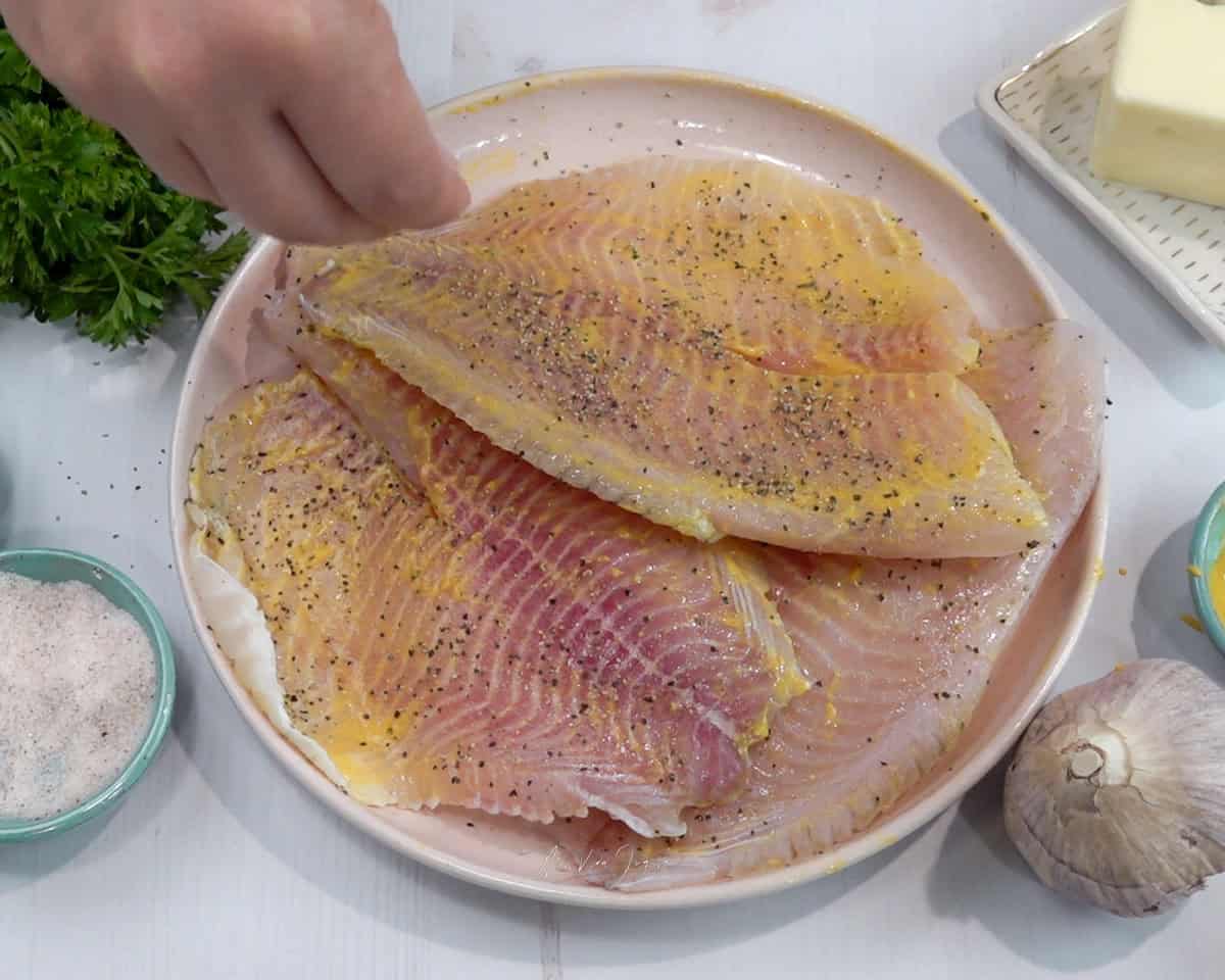 Una mano condimenta con mostaza sal y pimienta los filetes de tilapia colocados en un plato; alrededor se observan una cabeza de ajo, una tacita con sal, un manojo de perejil y mantequilla.