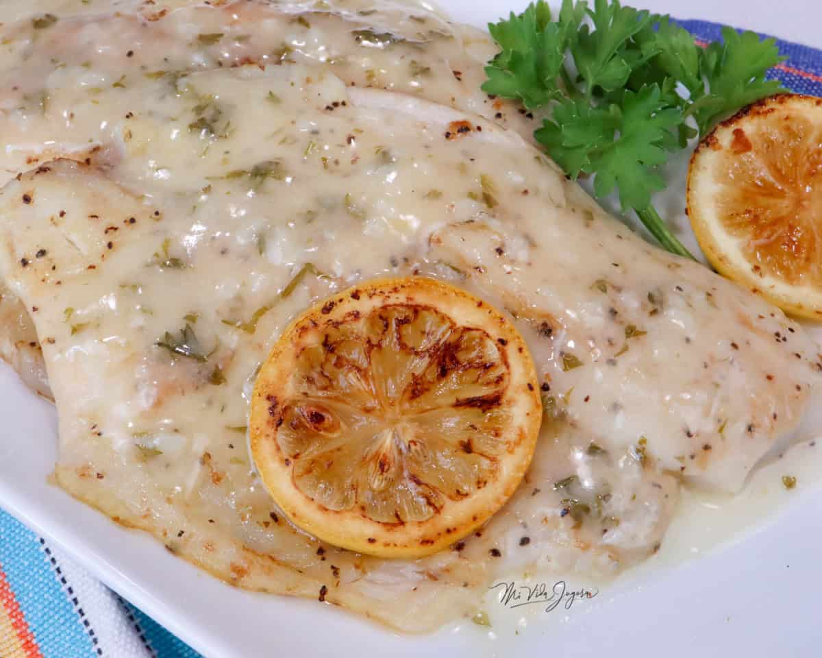 Un filete de tilapia cubierto con salsa, decorado con una rodaja de limón por encima. A un lado se observa otra rodaja de limón y unas hojas de perejil fresco.