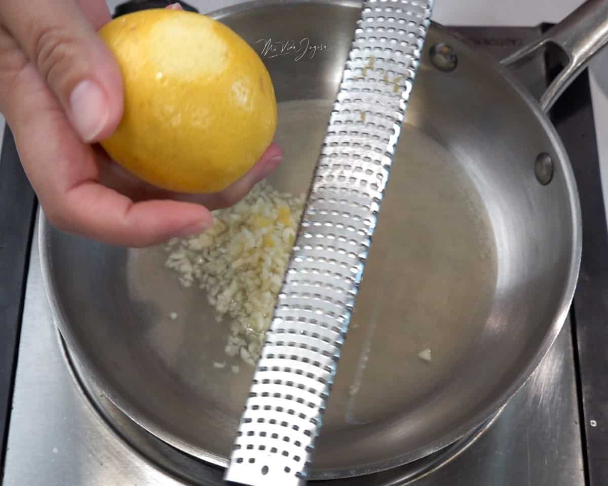 Una cacerola con jugo de limón y ajo en su interior; en primer plano, una mano sostiene un limón mientras la otra utiliza un zester para rallar su cáscara.