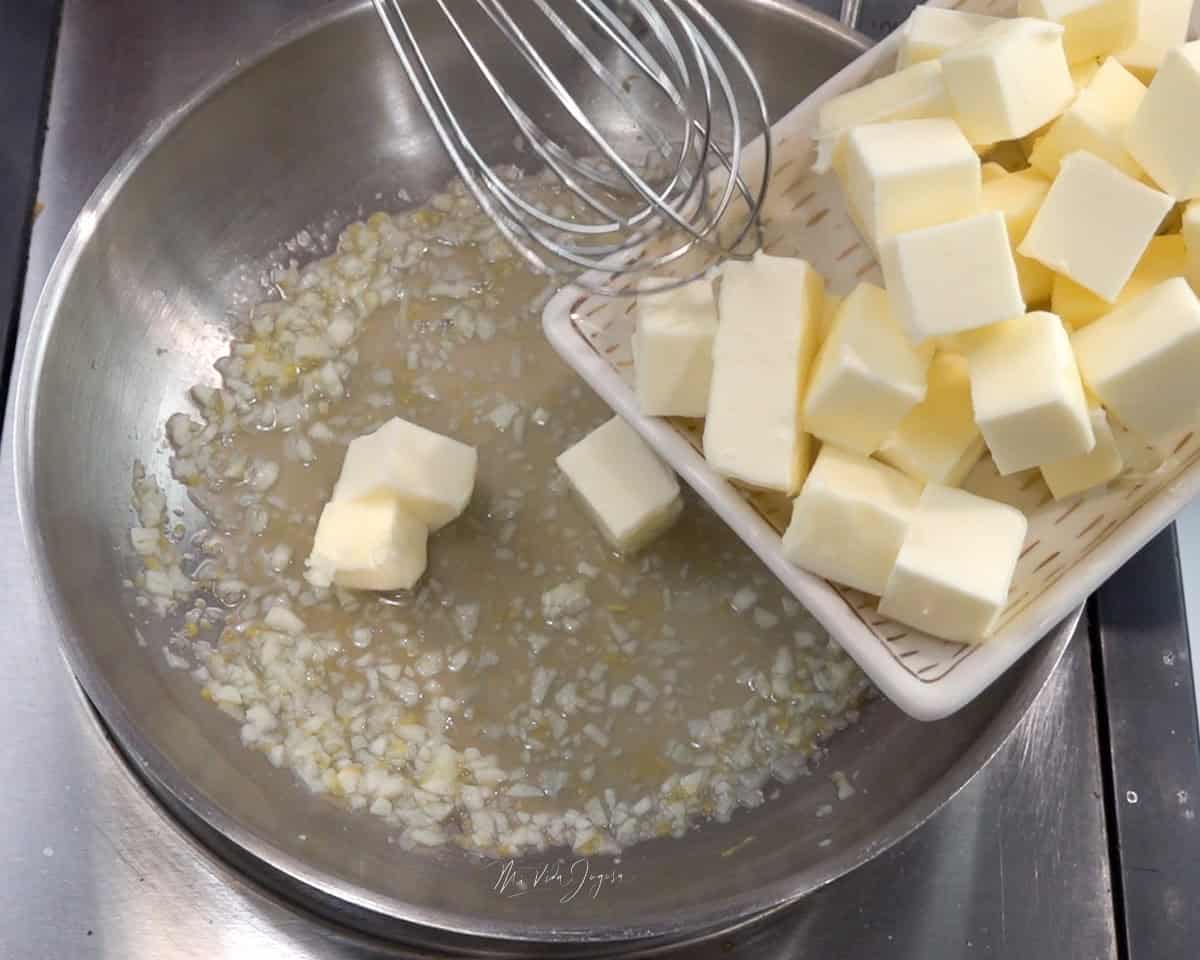 Un batidor de globo mezcla una cacerola con jugo de limón y ajo mientras se agregan pequeños cubos de mantequilla.