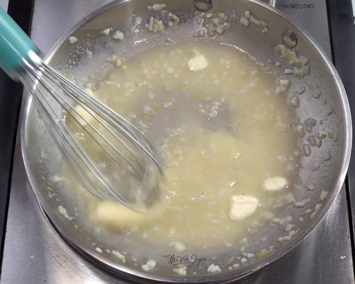 Una cacerola con cubos de mantequilla derritiéndose en jugo de limón y ajo, mientras la mezcla se bate con un batidor de globo.