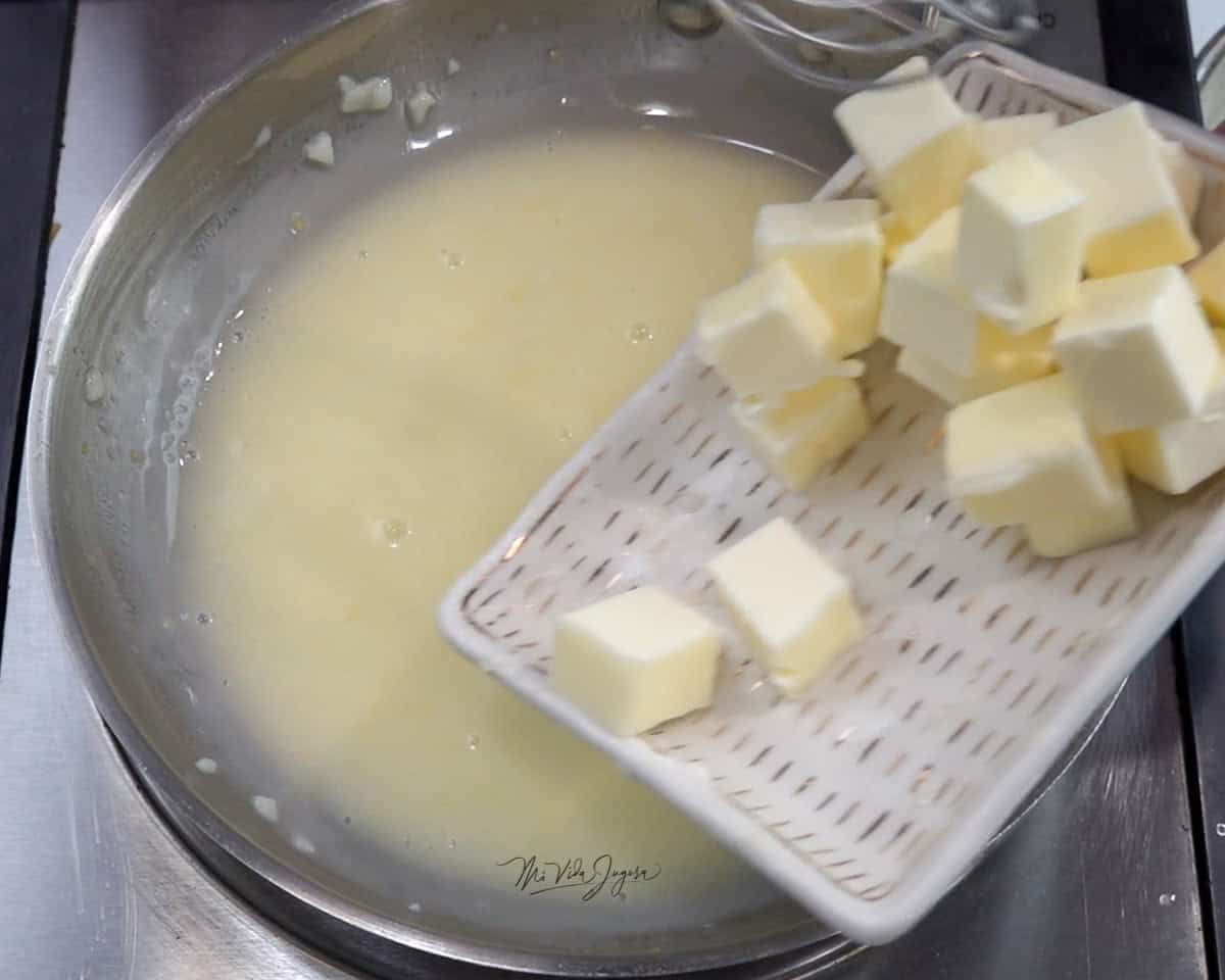 Una cacerola con una mezcla de mantequilla, limón y ajo, mientras por encima se observa un plato con pequeños cubos de mantequilla listos para agregarse.