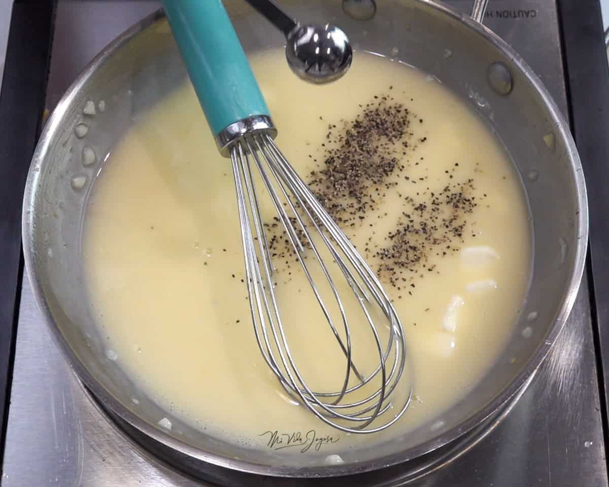 Una cacerola con la mezcla de la salsa de mantequilla, limón y ajo; un batidor de globo dentro de la cacerola y una cuchara agregando pimienta a la mezcla.