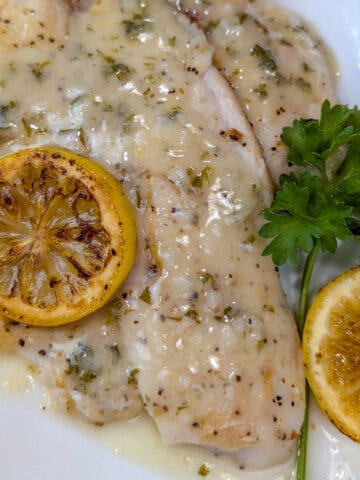 En un plato, un filete de tilapia bañado en salsa de mantequilla, decorado con una rodaja de limón encima; al lado, una hoja de perejil y otra rodaja de limón.