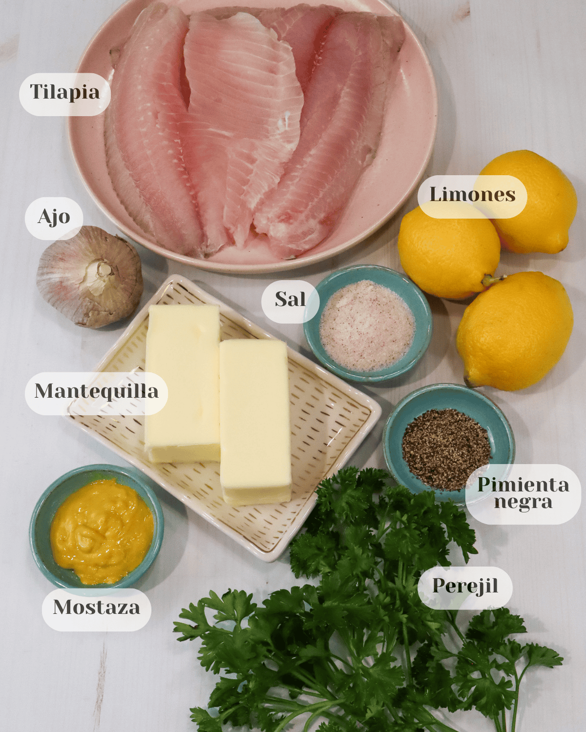 Sobre una superficie se encuentran los ingredientes de la receta: en un plato, varios filetes de tilapia; una cabeza de ajo, tres limones, una tacita con sal y pimienta, un plato con dos barras de mantequilla, una tacita de mostaza y un manojo de perejil.