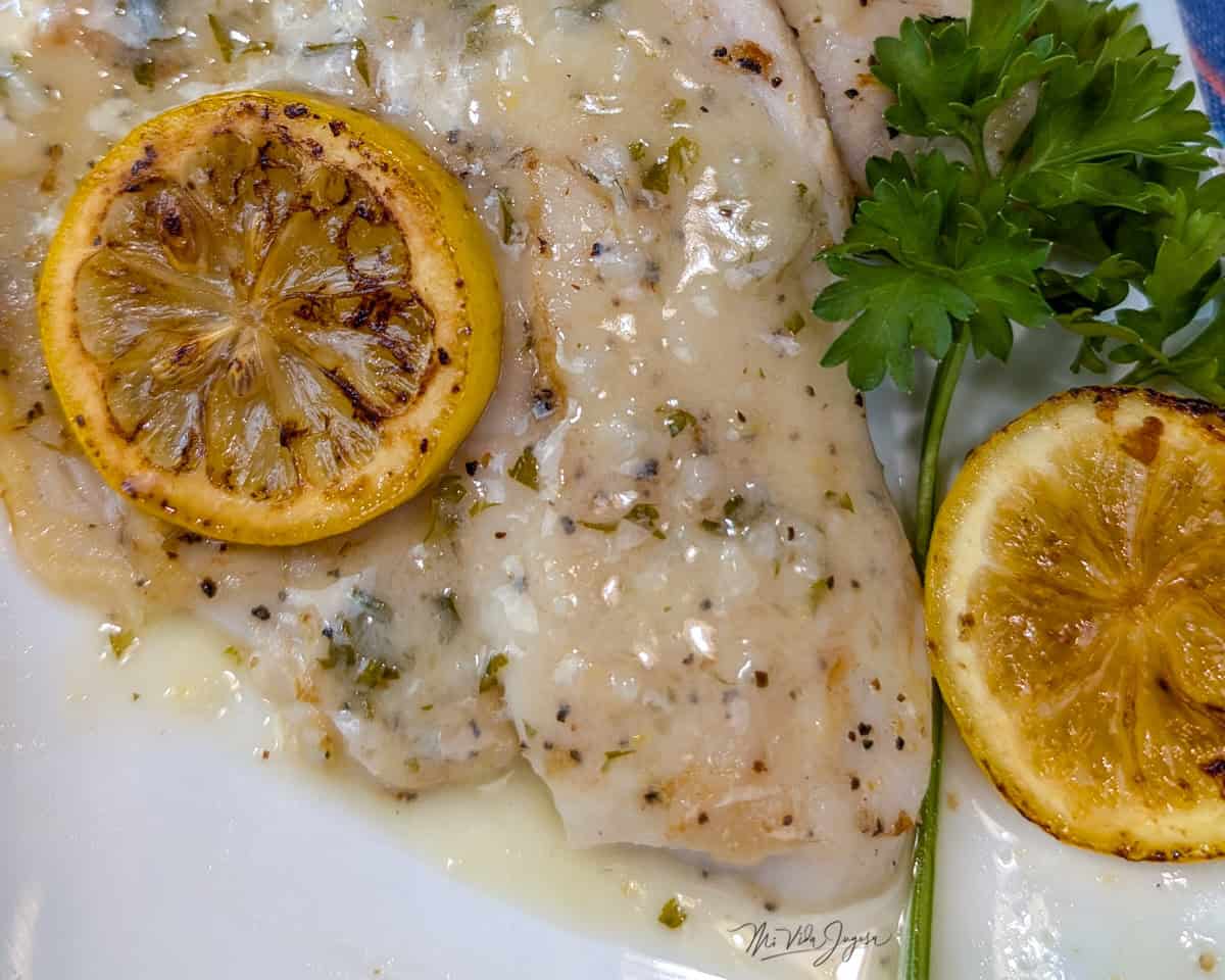 Un filete de tilapia cocinado y bañado en salsa, decorado con una rodaja de limón por encima; a un lado se observan una rodaja de limón y una rama de perejil.