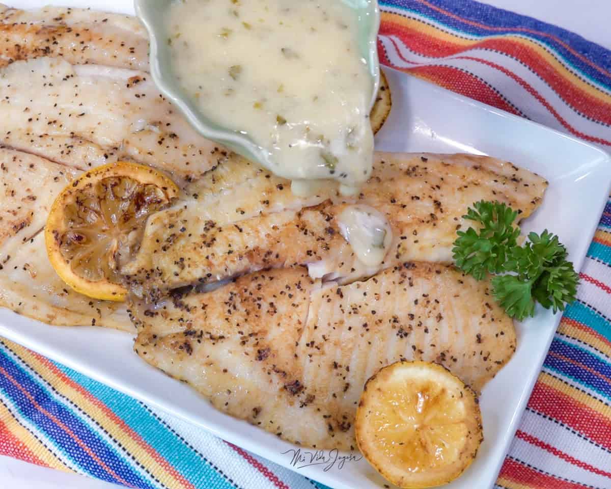 Un plato blanco cuadrado que contiene 2 tilapias cocinadas y dos rodajas de limon acompañando el platillo, mientras una tacita pequeña agrega salsa de mantequilla, limon y ajo a las tilapias.