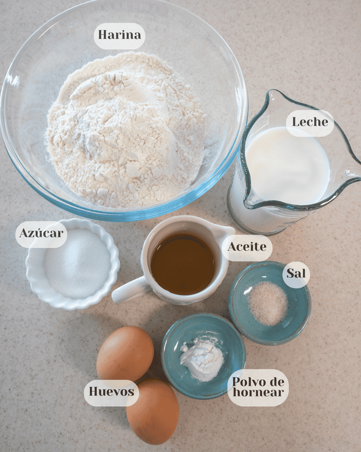 Un tazón con harina, un vaso medidor con leche, una tacita con azúcar, un recipiente con aceite, y otros con sal, polvo de hornear y dos huevos.