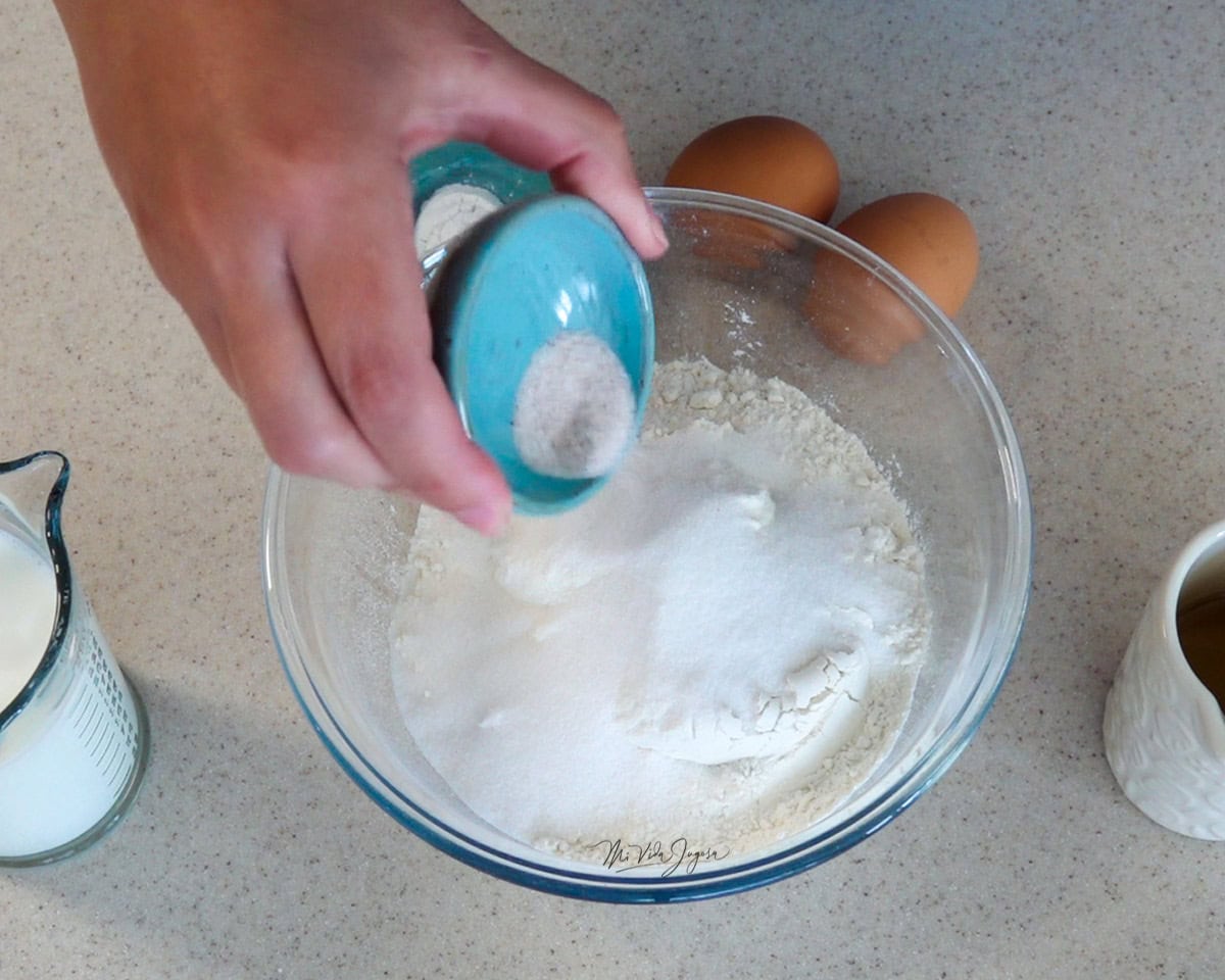 Un bowl con harina y azúcar, mientras una mano agrega la sal desde una tacita. Alrededor del tazón se ven unos huevos, una taza con aceite y otra con leche.