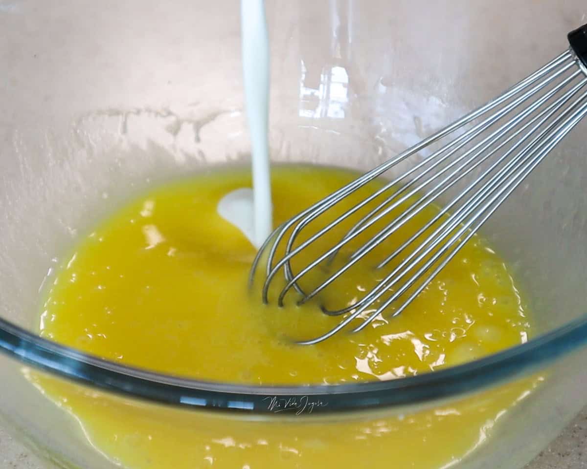 Un tazón con huevos ligeramente batidos, un batidor de globo y, al mismo tiempo, leche incorporándose a la mezcla.