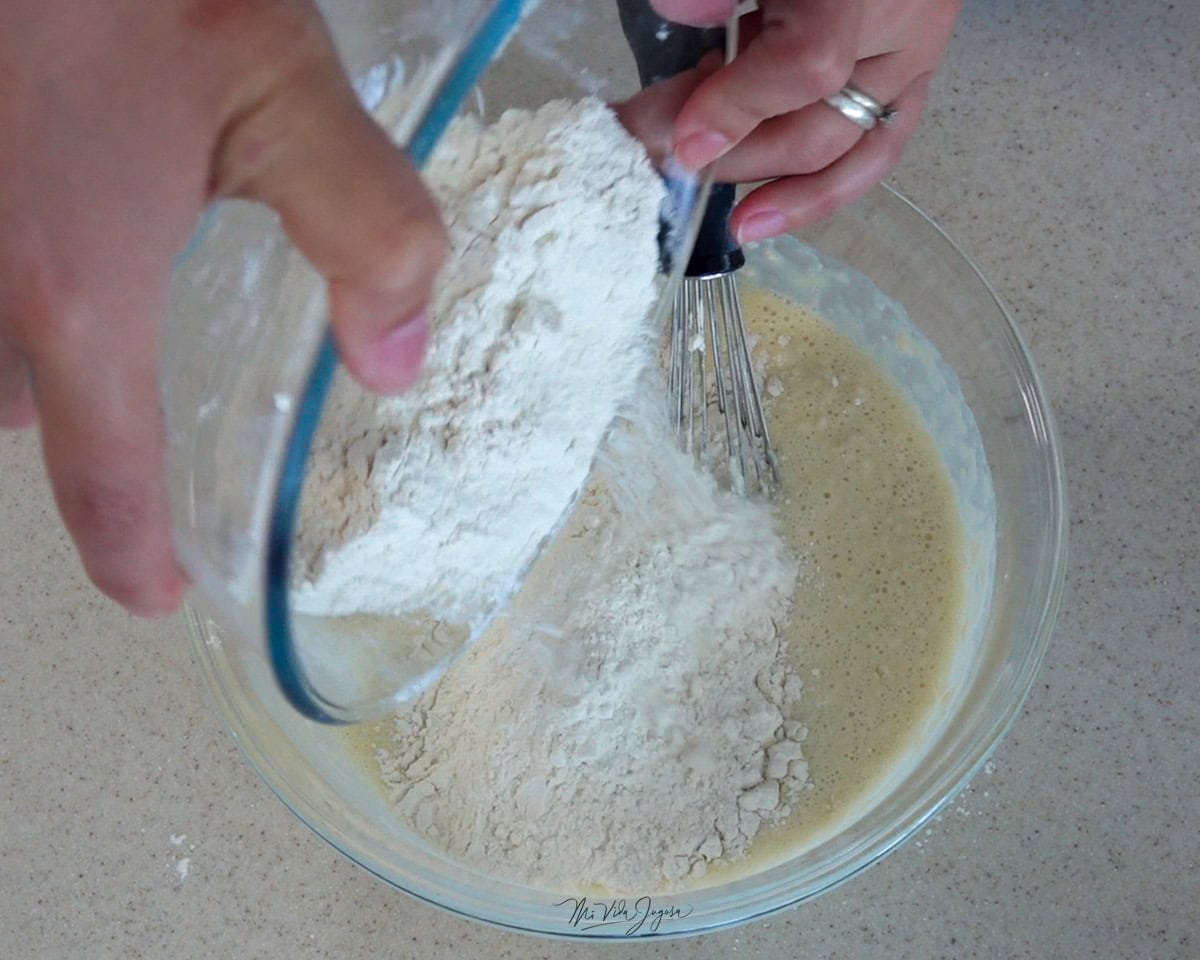 Un tazón con los ingredientes líquidos y un batidor de globo, en otro tazón los ingredientes secos siendo incorporados al tazón de los líquidos.