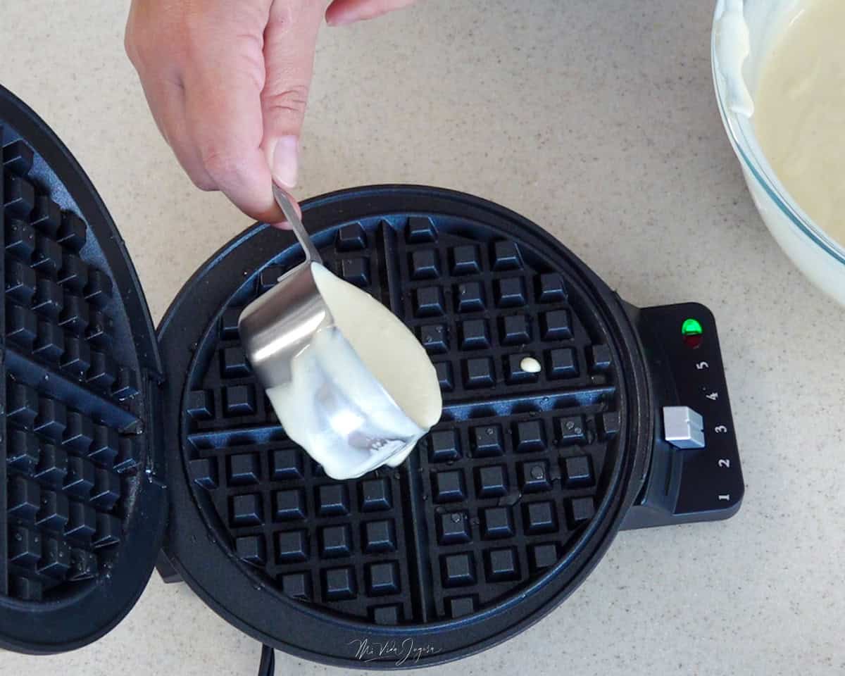 Una mano sostiene una taza con mezcla para waffles, vertiéndola cuidadosamente en la wafflera.