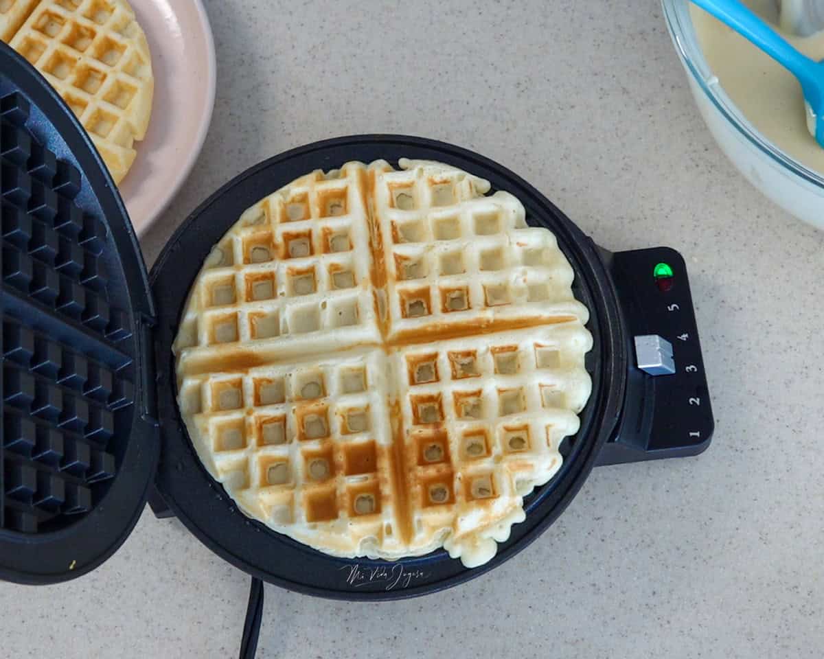 Un waffle cocinado en la máquina de waffles, mientras al lado se observa un plato con otro waffle y un tazón con la mezcla.
