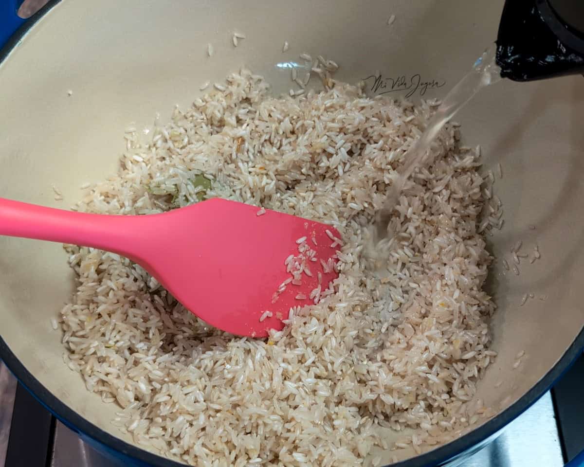 Una olla con una espátula dentro, que contiene granos de arroz crudos, mientras un pichel vierte agua sobre el arroz.