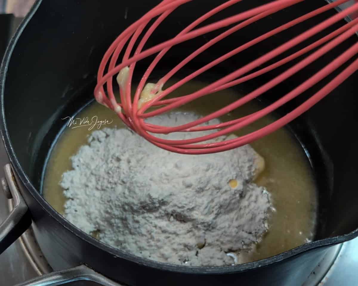 Una pequeña olla con mantequilla derretida y harina encima, mientras se introduce un batidor de globo.