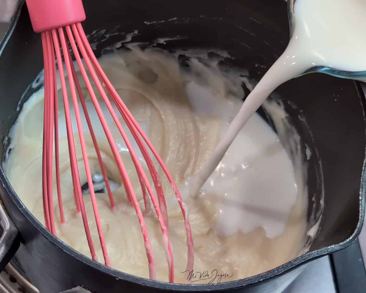 Una olla con una mezcla de mantequilla, harina y leche, mientras un pichel incorpora un chorrito de leche y un batidor de globo revuelve la mezcla.