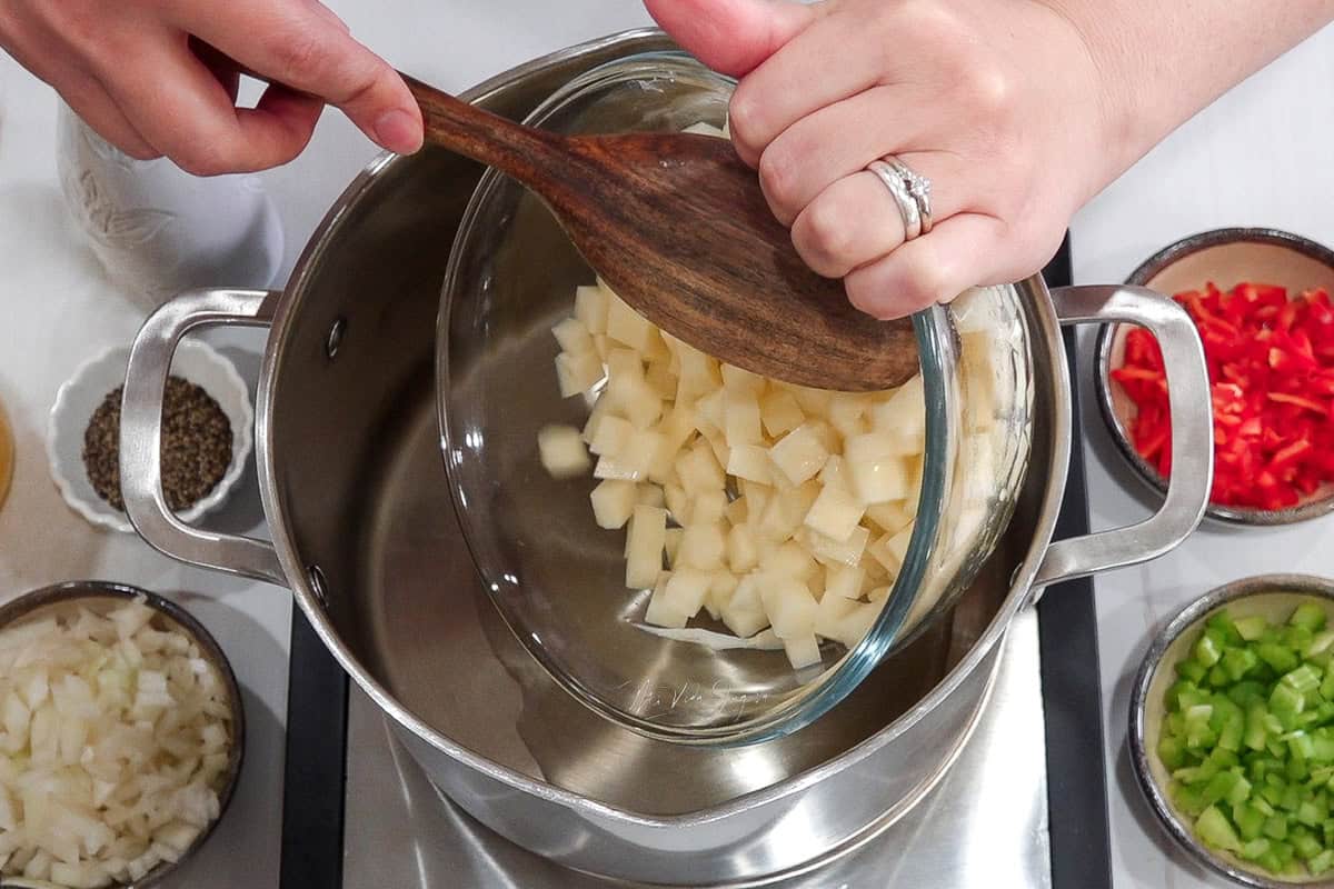 Una olla a la cual se le esta incorporando unos trocitos de papa que se encuentran en un bowl con ayuda de una cuchara de madera que esta siendo sostenida el boel y la cuchara por unas manos, al rededor de la olla se encuentran varias tacitas con cebolla, chile y apio y pimienta