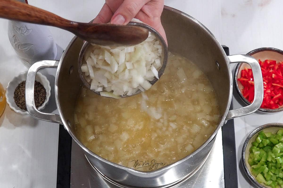 Una mano sostiene una tacita de cebolla que esta siendo incorporada a la olla con papas, al lado se observa una tacita con chile dulce y apio
