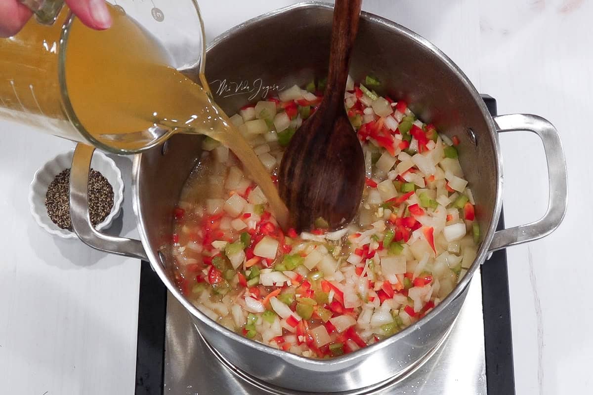 Una mano sostiene un pichel con caldo que se está vertiendo en una olla donde se están cocinando papas y otros vegetales.