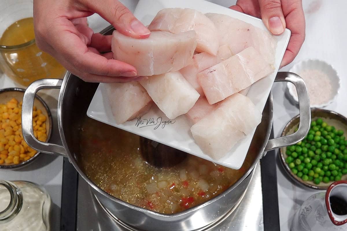 Un plato conteniendo pedazos de bacalao crudo, mientras una mano agarra un pedacito para incorporarlo a la olla que se encuentra debajo del plato, al lado se ven los guisantes y el maiz dulce en tacitas, y el caldo, la sal y el aceite
