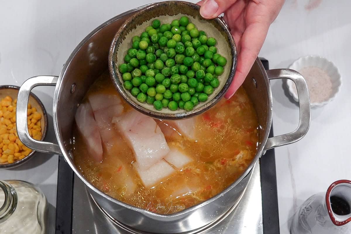 Una taza sostenida por iuna mano que contiene guisantes que encuentra por encima de la olla que contiene el bacalao y los otros ingredientes