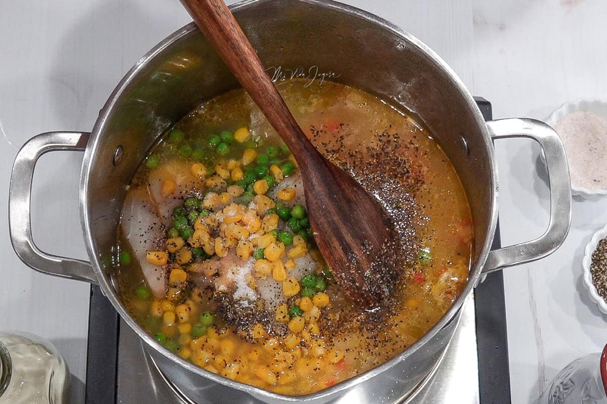 Una olla con la sopa en preparación, donde se observan guisantes y maíz dulce en la superficie, y debajo el resto de los ingredientes. En el centro hay una cuchara de madera lista para mezclar.