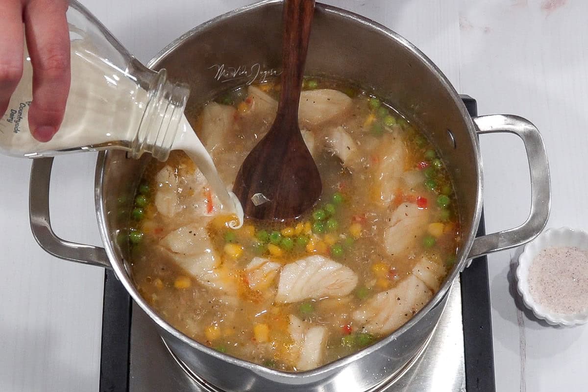 Una olla conteniendo unos guisantes, maiz dulce por encima y por debajo el resto de ingredeintes de la sopa, con una cuchara de madera en el medio
Una olla con la sopa en preparación, donde se observan guisantes y maíz dulce en la superficie, y debajo el resto de los ingredientes. En el centro hay una cuchara de madera lista para mezclar.
Una mano incorpora la crema dulce que se encuentra en un pichelito a la mezcla de la sopa de bacalao a la olla
Una mano vierte crema dulce desde un pequeño pichel sobre la sopa de bacalao que se está cocinando en la olla.