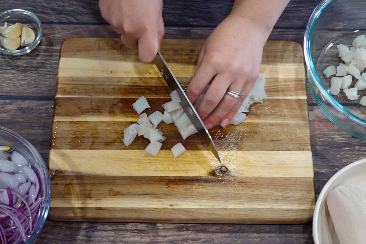 Sobre una tabla de picar, una mano sostiene las tiras de pescado mientras la otra, con un cuchillo, corta en sentido contrario para formar cubos; al lado hay un tazón con julianas de cebolla en agua con hielo, unos ajos y un tazón con cubos de pescado.