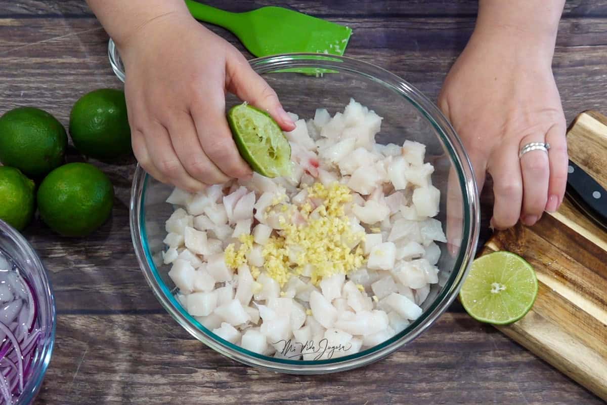 Una mano exprime un limón sobre un tazón con pescado y ajo; al lado hay unos limones y una espátula.