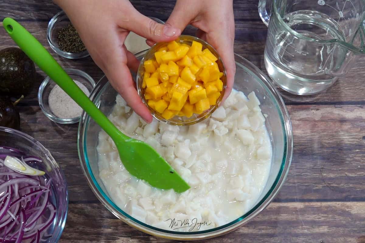 Un tazón con una espátula dentro, encima, un par de manos sostiene una tacita con cubitos de mango. Alrededor del tazón hay una taza con cebolla en julianas, sal, pimienta, maracuyá y una taza con Sprite.