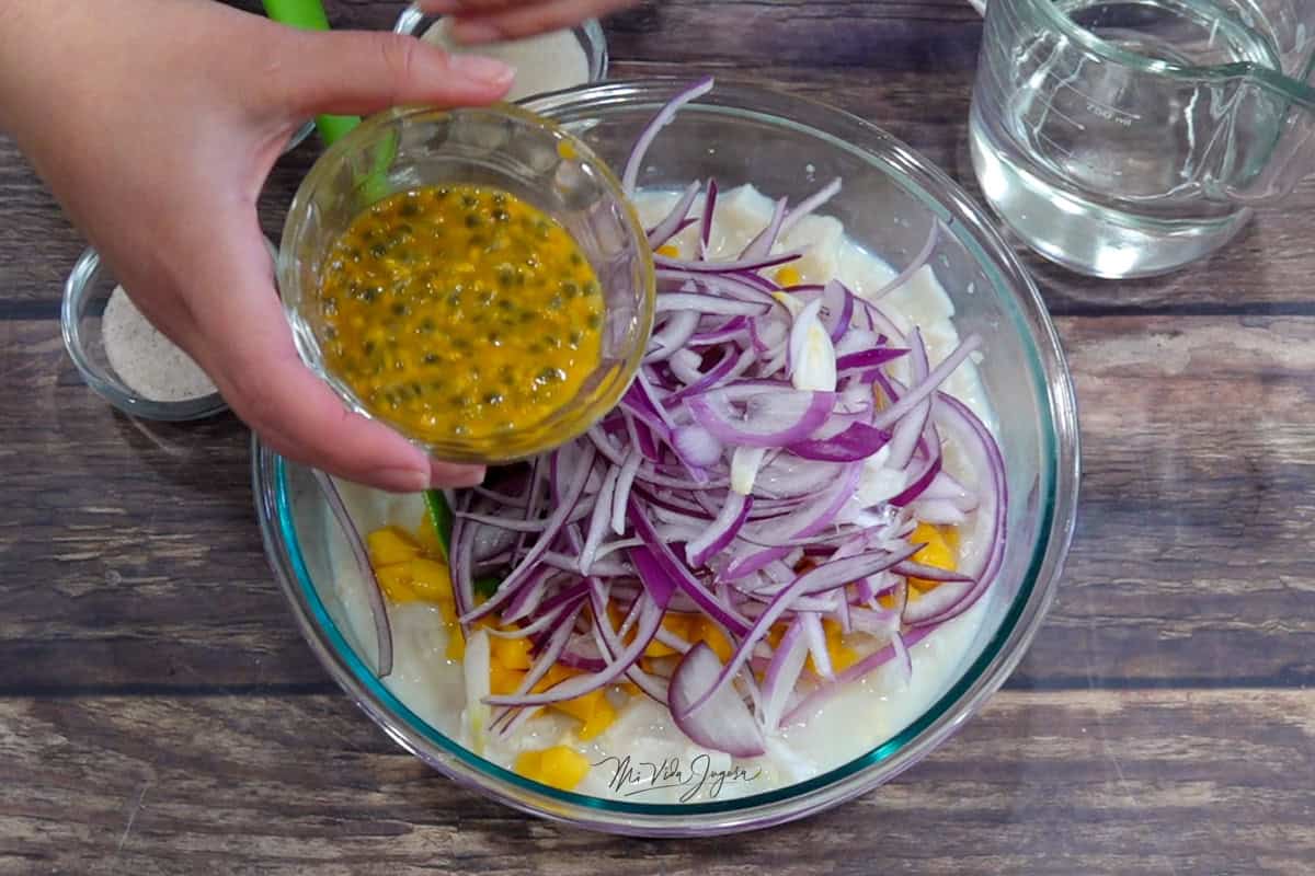 Un tazón con cubitos de pescado, cebolla en juliana y mango; por encima, una mano sostiene una tacita con pulpa y semillas de maracuyá lista para incorporarse. Al lado hay una taza con Sprite, sal y azúcar.