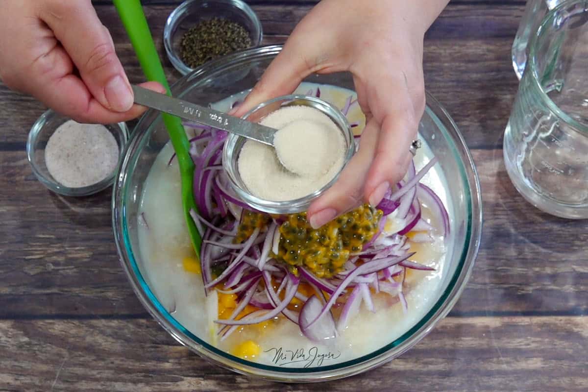 Un tazón con pescado, mango, cebolla y maracuyá; por encima, una tacita con azúcar mientras una cuchara medidora la incorpora. Al lado se ven pimienta, sal y Sprite en una taza de medida.