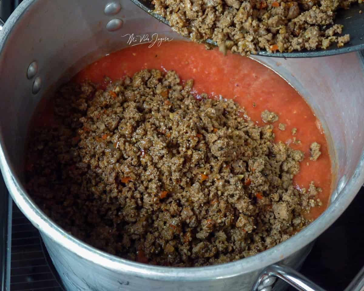 Una olla con salsa de tomate, mientras se agrega la carne molida poco a poco para mezclarla con la salsa.