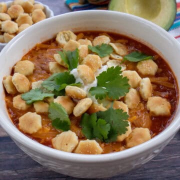 Una taza de chili con carne servida con queso rallado, natilla, culantro fresco y mini galletitas por encima. A un lado se observa un mantel decorativo, medio aguacate y una tacita con mini galletas.