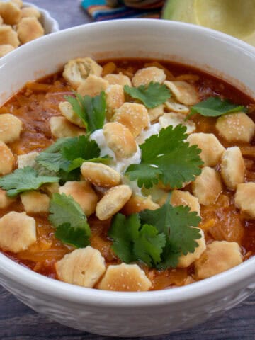 Una taza de chili con carne servida con queso rallado, natilla, culantro fresco y mini galletitas por encima. A un lado se observa un mantel decorativo, medio aguacate y una tacita con mini galletas.