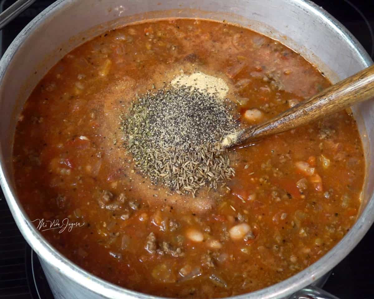 Una olla con chili con carne y condimentos por encima, mientras una cuchara se encuentra dentro de la mezcla.