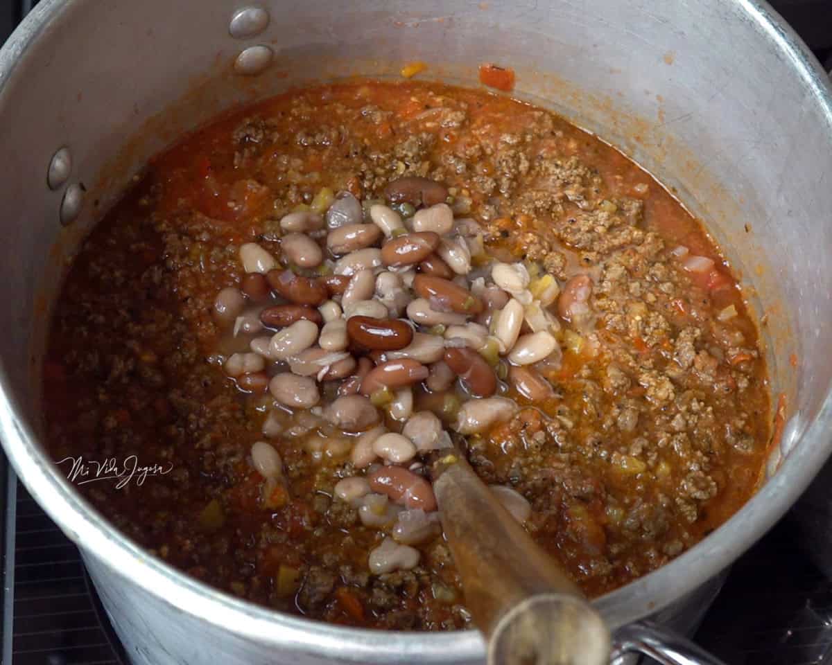 Una olla con carne molida en salsa de tomate y frijoles enteros por encima. Dentro de la olla se observa una cuchara lista para mezclar los ingredientes.