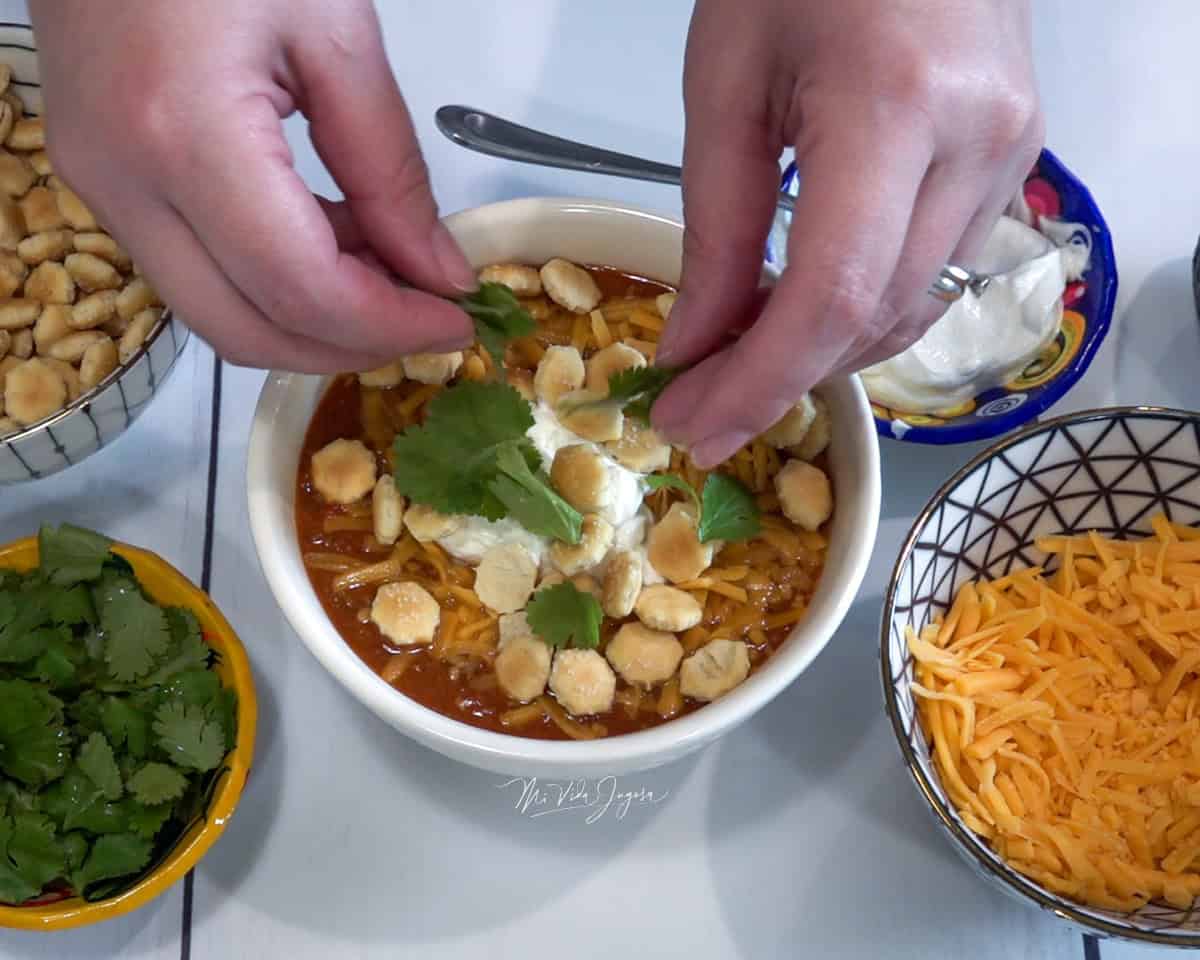 Una tacita de chili con carne servida con queso rallado, natilla y galletitas por encima, mientras dos manos agregan culantro fresco. A un lado se observan varias tacitas con galletitas, natilla, culantro y queso amarillo rallado.