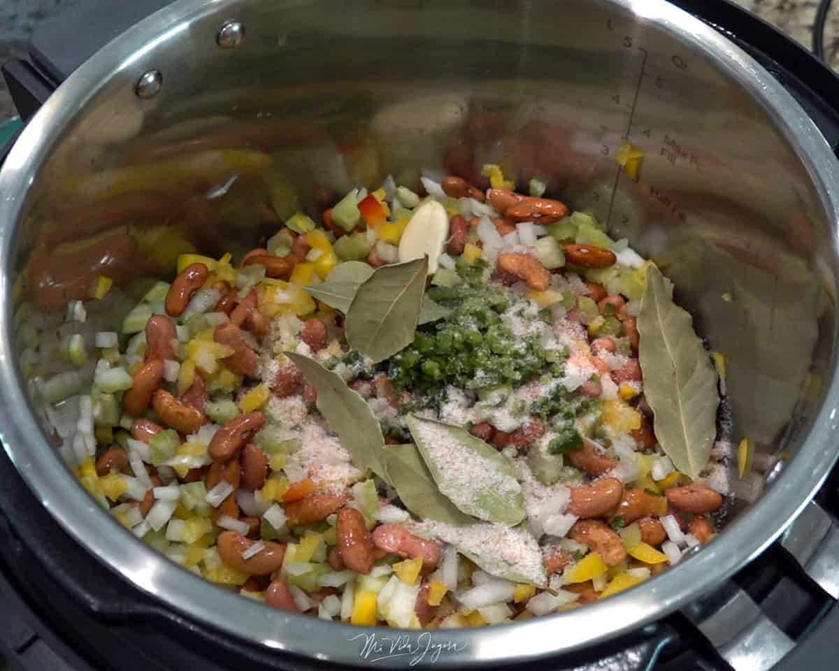 Una olla con frijoles, chile dulce y cebolla picada. Por encima se observan unas hojas de laurel y sal, listas para comenzar la cocción.