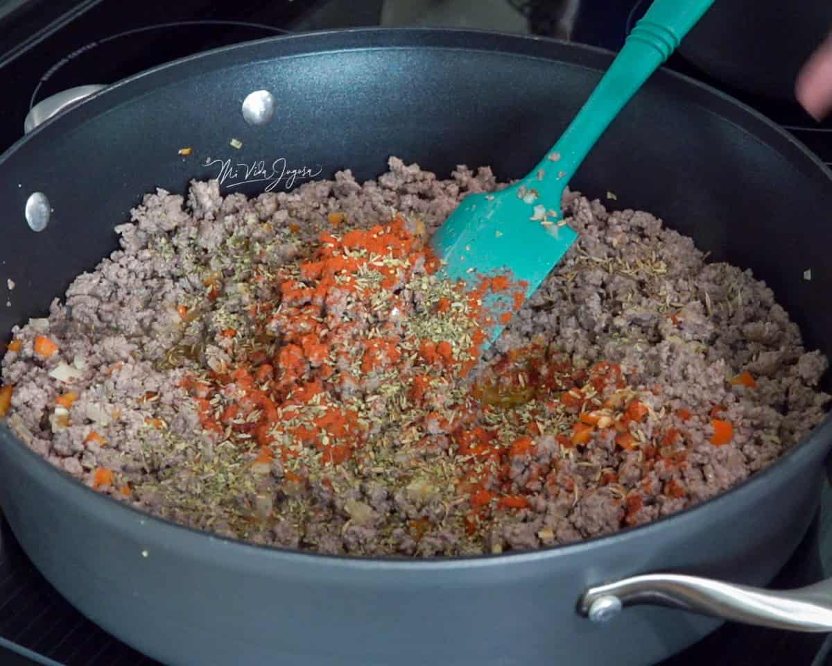 Un sartén con carne molida ya cocinada, cubierta con condimentos como paprika y sazón completo, mientras una espátula la mezcla para integrar mejor los sabores.