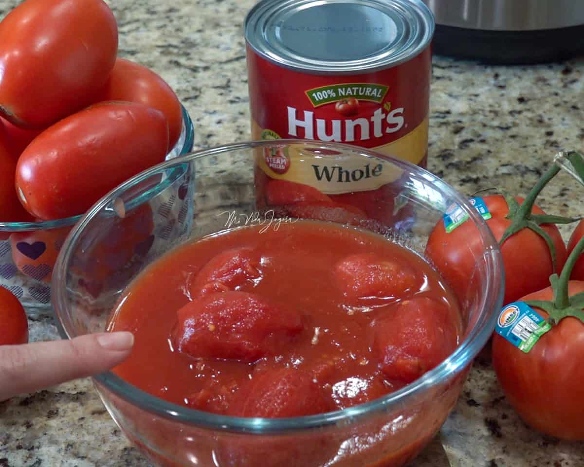 Un bowl de vidrio con salsa de tomates enteros. Detrás se observa una lata de tomates, un bowl con tomates naturales y, a un lado, unos tomates frescos en rama.
