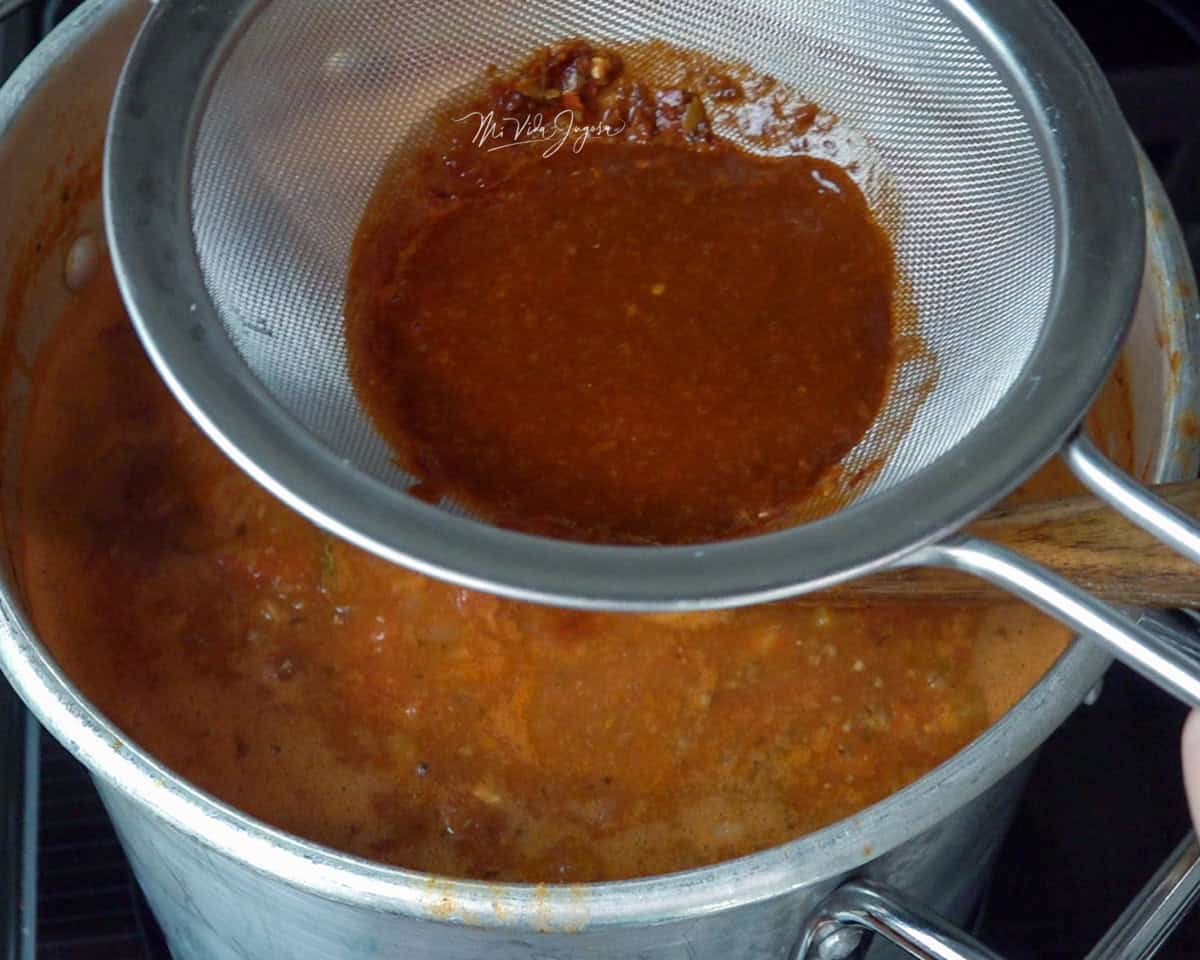 Una olla con chili con carne y un cucharón dentro de la mezcla. Por encima se observa un colador filtrando una salsa de chiles que se está incorporando poco a poco al chili.
