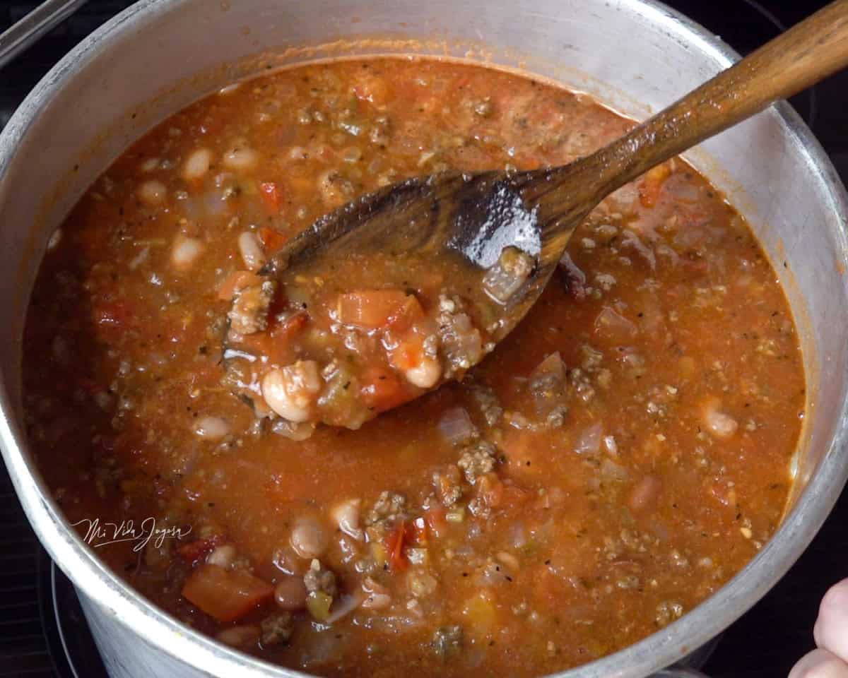 Una olla con chili con carne, mientras una cuchara se levanta ligeramente sobre la mezcla para mostrar la carne, los frijoles enteros y los pedacitos de tomate.