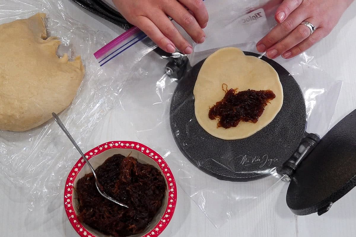 Una tortillera con un papel plástico en el centro sostiene un disco de masa aplanado y relleno de chiverre. Dos manos están a punto de cerrar la masa para formar la empanada; a un lado se observa una tacita con dulce de chiverre y un tenedor dentro, y al lado una bolita de masa lista para usar.