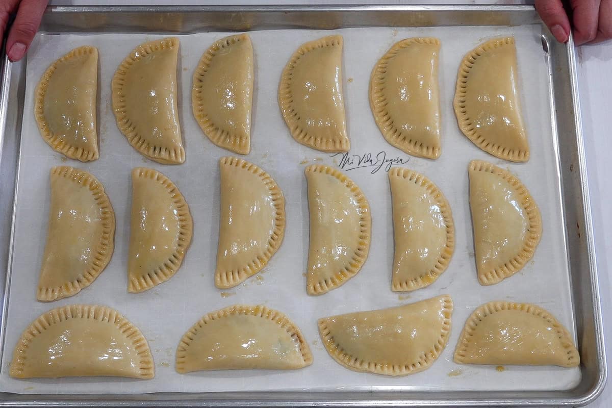 Una bandeja cubierta con papel pergamino y empanadas barnizadas con huevo, mientras dos manos sostienen la bandeja