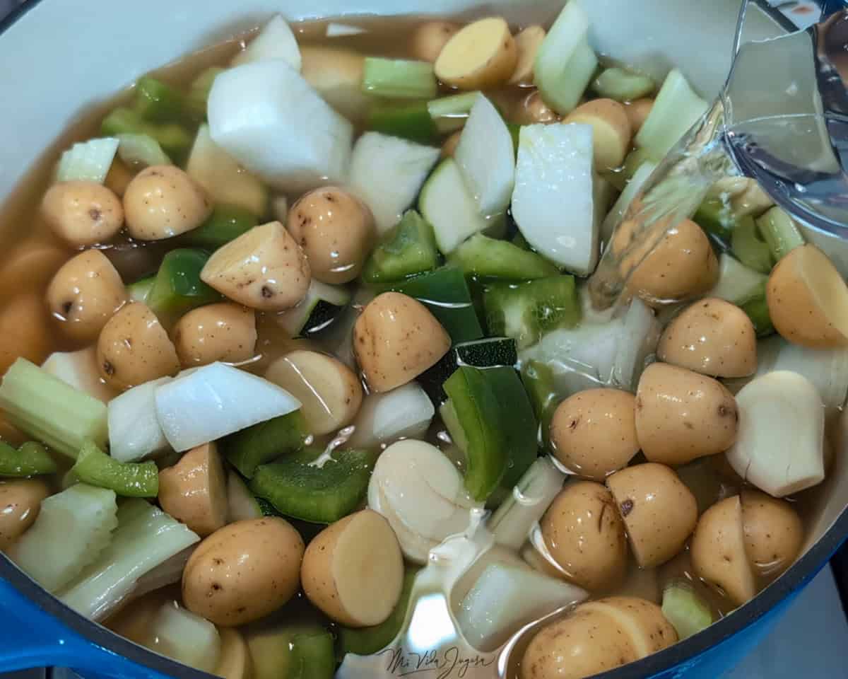 Una olla contiene pedazos de chayote, zucchini, papa, cebolla y chile verde, mientras un pichel vierte agua dentro.