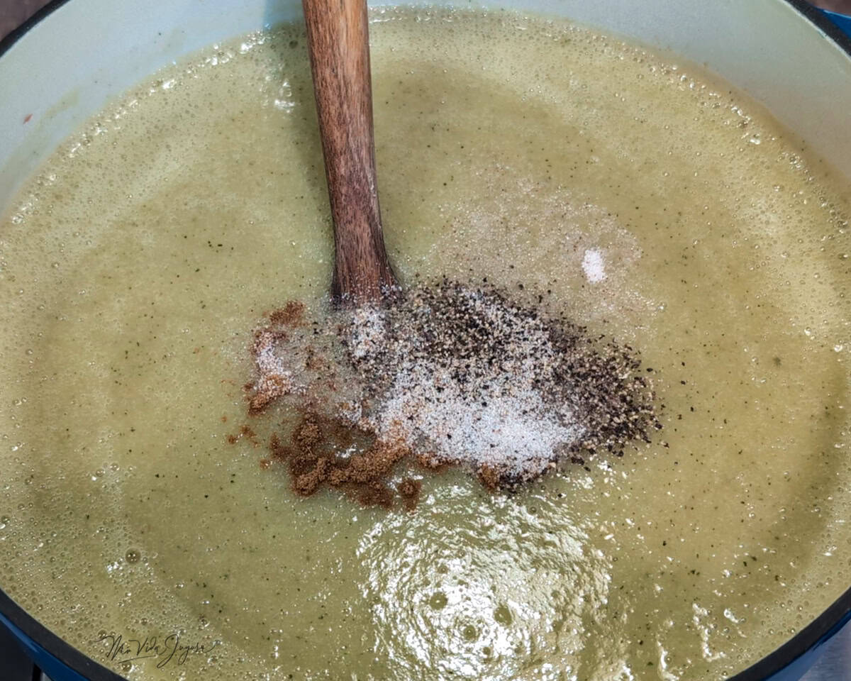 Una olla contiene crema de vegetales con una espátula de madera dentro. Encima de la crema se observan comino, pimienta y sal.