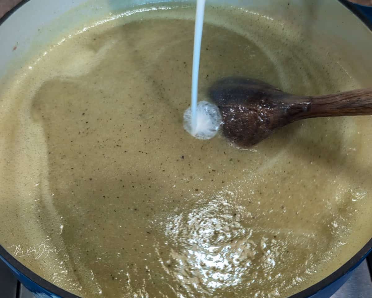 Una olla contiene crema de vegetales con una espátula dentro, mientras se agrega crema dulce.