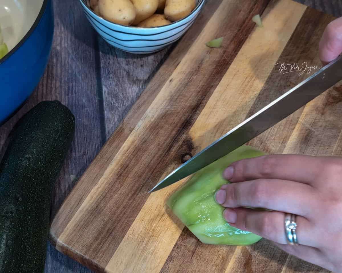 Sobre una tabla de picar, una mano sostiene medio chayote pelado, mientras la otra mano lo corta con un cuchillo. A un lado se observa un zucchini y varias tacitas con papas mini.