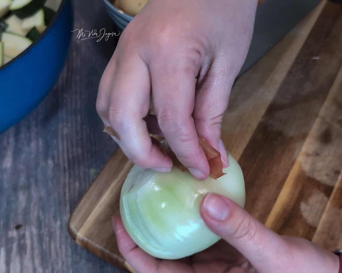 Una mano sostiene una cebolla mientras la otra retira la cáscara seca. Al fondo se visualiza una tabla de picar y una taza con pedacitos de vegetales.